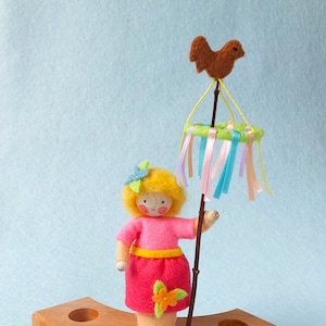 Pode incluir: Uma boneca de feltro com cabelo loiro e um vestido rosa segurando um mastro de maio com um pássaro marrom no topo. O mastro de maio tem fitas coloridas e uma coroa verde. A boneca está em pé sobre uma base de madeira.