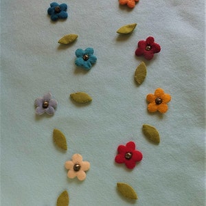 Puede incluir: Un fondo azul claro con flores de fieltro en varios colores, incluyendo azul, rojo, naranja, amarillo y rosa. Las flores están dispersas por el fondo y tienen un diseño simple de cinco pétalos. Cada flor tiene un pequeño botón dorado en el centro.