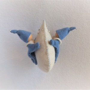 May include: A handmade felt Christmas decoration. The ornament is a light cream colour with a pointed shape, adorned with small silver beads. It features two figures with blue hats and mittens, and a blue cape.