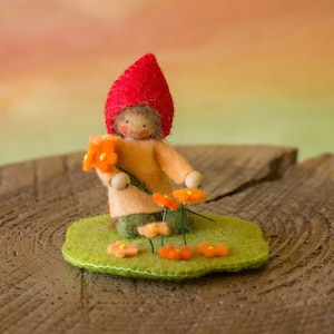 May include: A felt doll wearing a red hat and holding orange flowers. The doll is standing on a green felt base with more orange flowers.