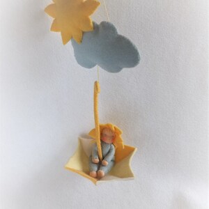 Op de afbeelding: Een vilt mobiel met een gele zon, een blauwe wolk en een kleine viltpop die in een gele paraplu zit.
