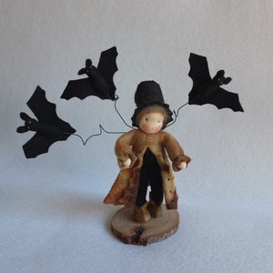 May include: A felt doll wearing a brown coat and a black top hat, holding three black felt bats with wire wings.
