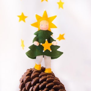 May include: A felt doll dressed as a Christmas tree with a yellow star on its head and a yellow star in its hand. The doll is standing on a brown pine cone.