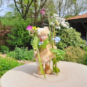 Könnte beinhalten: Eine kleine Holzpuppe mit einem hellbraunen Kleid und weißen Haaren hält einen Strauß aus rosa, blauen und weißen Blumen. Die Puppe steht auf einer Holzoberfläche.