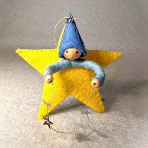 May include: A handmade felt star ornament with a whimsical star-shaped figure. The figure has a blue hat and arms, holding a string of silver stars. The star is yellow, and the figure's face is painted with simple details.