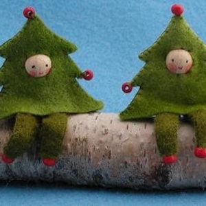 May include: Three felt Christmas tree ornaments, each with a small wooden face, are sitting on a white birch log. The ornaments are green with red berries and are wearing a green felt outfit.