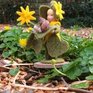 May include: A felt doll with yellow hair and a yellow flower crown is holding a green leaf with brown snails. The doll is standing on a wooden base in a garden setting with yellow flowers and green leaves.