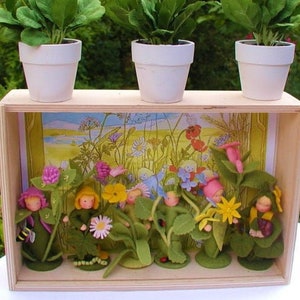 May include: A wooden box with a miniature garden scene inside. The box has three white flower pots with small daisy plants on top. Inside the box, there are miniature felt figures of people standing in a field of flowers. The flowers are yellow, pink, and purple.