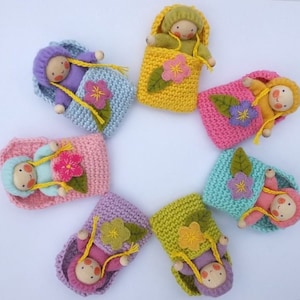 Op de afbeelding: Handgemaakte poppen in kleurrijke gehaakte slaapzakken. Elke pop heeft een houten hoofd, garen haar en is genesteld in een tas versierd met een vilten bloem en bladeren. De tassen zijn in roze, geel, blauw en paars.