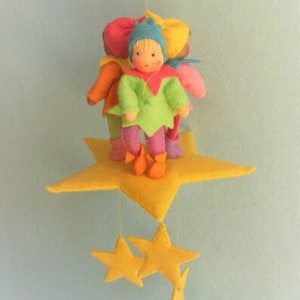 May include: A colourful mobile with felt dolls in various outfits, standing on a large yellow star. Smaller yellow stars hang below. The dolls are dressed in green, orange, pink, and blue. The mobile is set against a light blue backdrop.