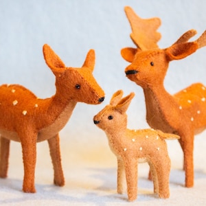 May include: Three felt deer figurines in varying sizes. The deer are orange with white spots and black eyes. The largest deer has antlers. The figurines are standing on a white surface.