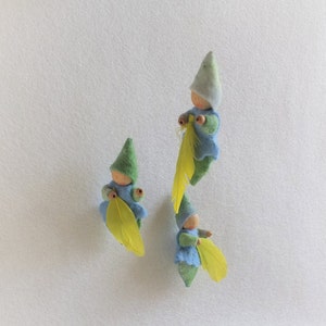 Op de afbeelding: Drie vilten dwergpoppen met groene hoeden en blauwe shirts die gele bladeren vasthouden. De dwergen staan op een witte achtergrond.