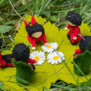 May include: Five felt ladybug dolls are gathered around a green felt leaf with white felt daisies. The ladybugs are red with black spots and black heads.