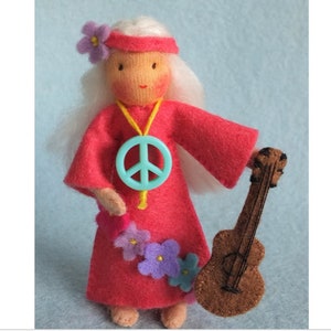 Op de afbeelding: Een vilten pop met lang wit haar, die een rode jurk draagt met een vredesketting en een bloemenkrans. De pop houdt een bruine vilten gitaar vast.