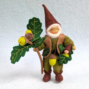 May include: A handcrafted felt gnome doll, dressed in olive green and brown, holding a branch with felt acorns and leaves. The gnome has a pointed brown hat and a white beard. The doll is approximately 6 inches tall.