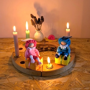 Może przedstawiać: Drewniany świecznik z zapalonymi świecami i dwiema filcowymi lalkami. Lalki ubrane są w różowe i niebieskie stroje i siedzą na żółtych klockach. Obecna jest również mała waza z suszonymi kwiatami i dekoracja ślimaka.