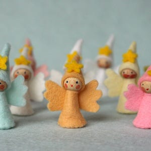 Könnte beinhalten: Eine Gruppe von acht Filzpuppen mit Flügeln und gelben Stern-Akzenten. Die Puppen sind in verschiedenen Farben gekleidet, darunter Blau, Rosa, Weiß und Orange.