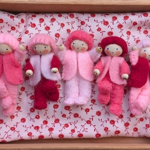 May include: Five handmade wooden dolls dressed in pink and red felt clothing. Each doll has a round wooden head with painted features and a fluffy pink hat. They are arranged in a wooden box on a floral patterned fabric.