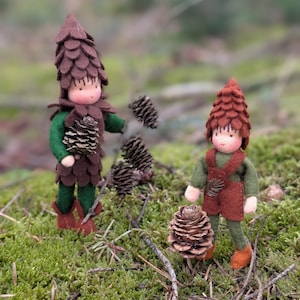 Può includere: Due bambole fatte a mano in feltro, vestite come creature della foresta, in piedi su un letto di muschio verde. Una bambola indossa un cappello marrone a forma di cono e una tunica abbinata, con delle pigne. L'altra bambola ha un cappello simile e un gilet rosso.