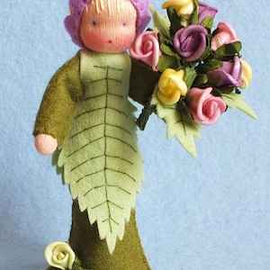 May include: A felt doll with a purple and yellow flower hat holding a bouquet of pink, purple, and yellow felt flowers. The doll is wearing a green felt dress with a leaf design.
