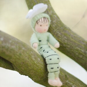 Puede incluir: Una mu&ntilde;eca de fieltro verde y blanco con una flor en la cabeza est&aacute; sentada en una rama de &aacute;rbol. La mu&ntilde;eca est&aacute; vestida como una oruga con puntos negros en su cuerpo.