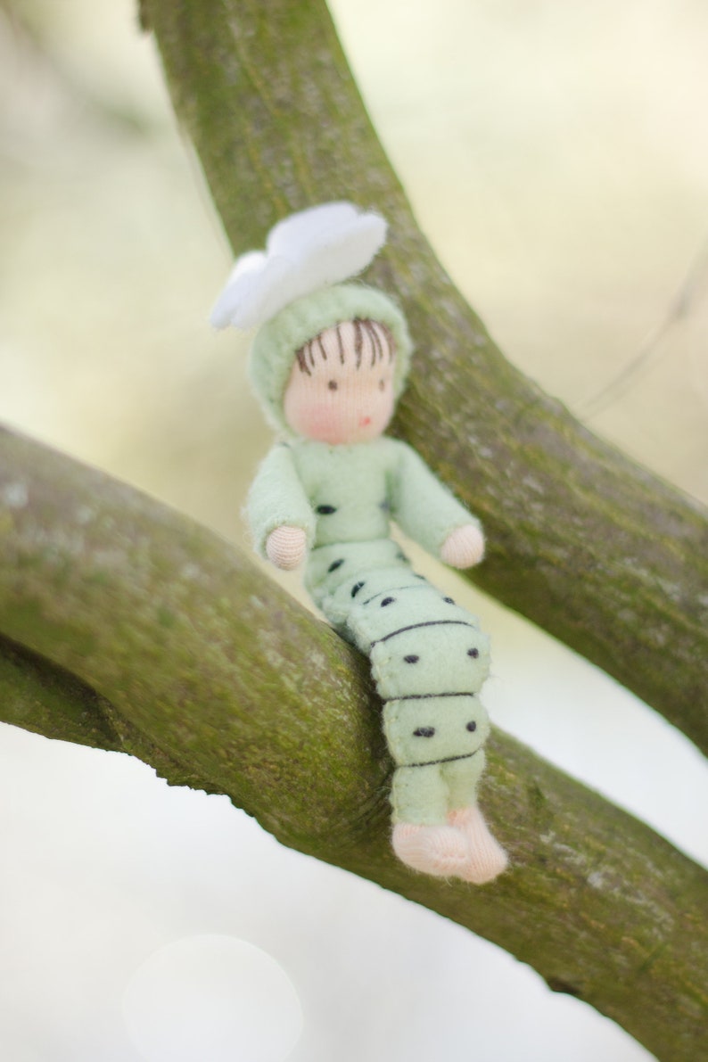 Puede incluir: Una mu&ntilde;eca de fieltro verde y blanco con una flor en la cabeza est&aacute; sentada en una rama de &aacute;rbol. La mu&ntilde;eca est&aacute; vestida como una oruga con puntos negros en su cuerpo.