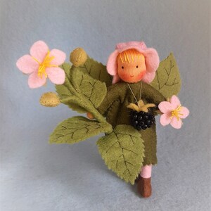 May include: A felt doll with blonde hair wearing a pink hat and green dress, holding a blackberry and standing in front of a felt flower bush with pink flowers and green leaves.