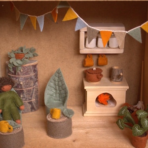 Op de afbeelding: Een miniatuur poppenhuis scène met een vilten pop, een klein houten fornuis en diverse vilten meubels en decoraties. De scène is versierd met een vilten slinger van vlaggen.