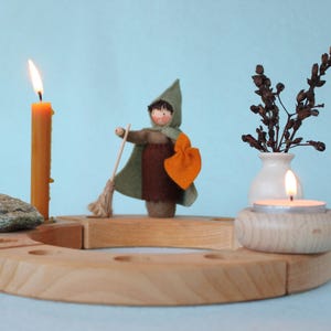 May include: A wooden advent calendar with four candle holders. The calendar features a small doll holding a broom and a vase with dried flowers. Two candles are lit.