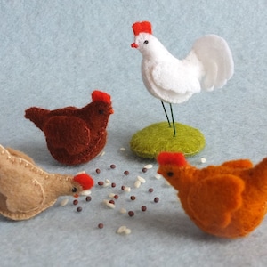 May include: A collection of handmade felt chicken figurines in various colors. The set includes a white rooster on a green base, and three hens in brown, tan, and orange, with red combs. Scattered grains add a touch of realism.