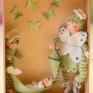 Puede incluir: Una caja de madera con un interior de fieltro verde. Tres mu&ntilde;ecas de fieltro est&aacute;n dentro de la caja. Una mu&ntilde;eca sostiene una almohada de fieltro verde. Otra mu&ntilde;eca est&aacute; acostada en una vaina de guisante de fieltro verde. La tercera mu&ntilde;eca est&aacute; de pie con una mariposa de fieltro blanco en el pecho.