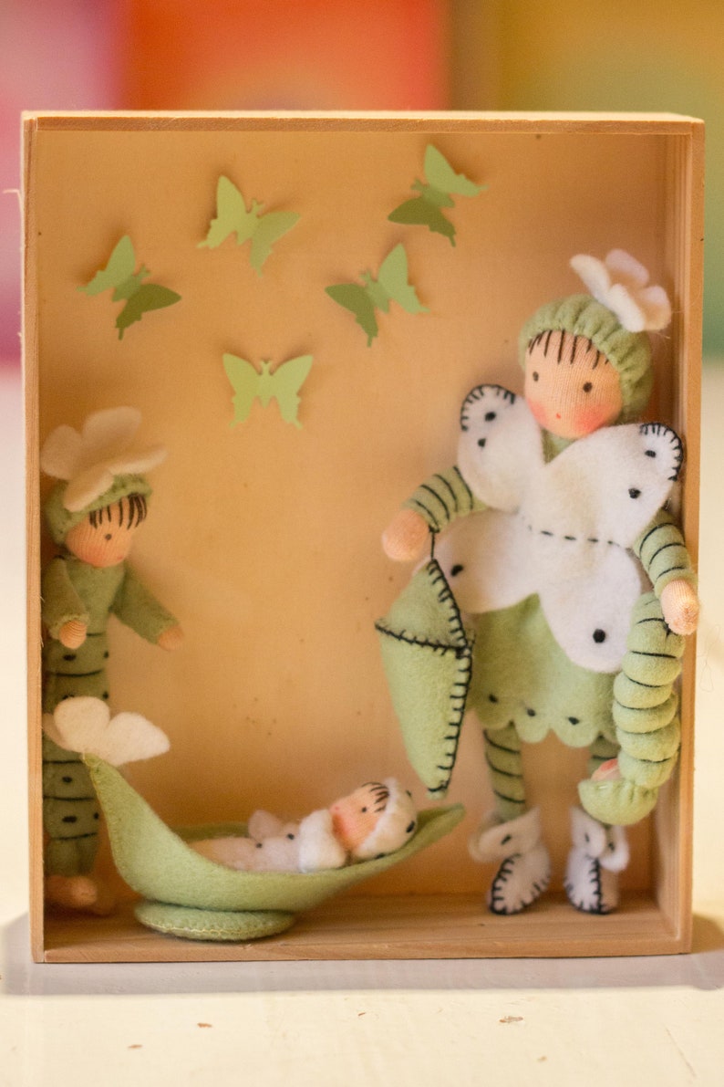 Puede incluir: Una caja de madera con un interior de fieltro verde. Tres mu&ntilde;ecas de fieltro est&aacute;n dentro de la caja. Una mu&ntilde;eca sostiene una almohada de fieltro verde. Otra mu&ntilde;eca est&aacute; acostada en una vaina de guisante de fieltro verde. La tercera mu&ntilde;eca est&aacute; de pie con una mariposa de fieltro blanco en el pecho.