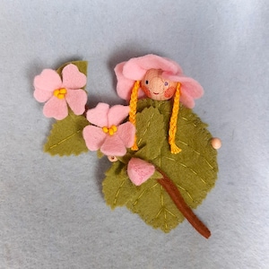 May include: A felt flower fairy doll with pink flowers and green leaves. The doll has a wooden head with a painted face and yellow yarn hair.