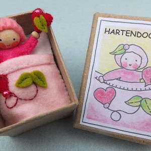 May include: A handcrafted wooden doll in a pink felt bed within a small cardboard box. The doll is wearing a pink hat and holding a red heart. The adjacent box features a drawing of a similar doll with the text "HARTENDOOSJE".