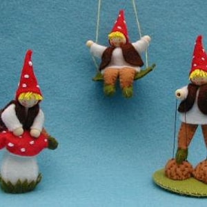Puede incluir: Tres muñecas de gnomo de fieltro, cada una con una actividad diferente. Un gnomo está sentado en un hongo rojo y blanco, otro está columpiándose en una hoja verde y el tercero está de pie sobre dos troncos marrones.