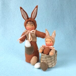 Puede incluir: Dos muñecas de conejo de fieltro, una más grande que la otra, sostienen huevos de Pascua. La coneja más grande lleva una cinta rosa alrededor del cuello. La coneja más pequeña está sentada en una cesta de mimbre.