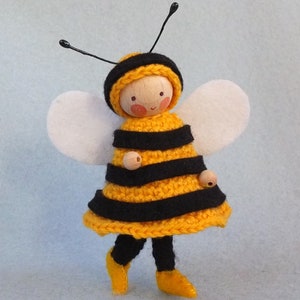 May include: A crocheted bee doll with black and yellow stripes, a wooden head, and white felt wings. The doll is wearing yellow shoes and has a black and yellow striped hat.