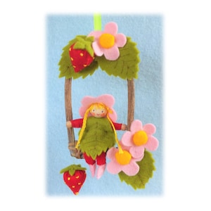 May include: A felt doll with blonde hair and a pink dress is sitting on a swing made of twigs and felt leaves. The swing is decorated with felt strawberries and pink flowers.