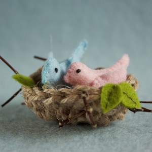 May include: Two felt birds, one blue and one pink, are nestled in a small woven nest made of twine. The nest is decorated with green felt leaves and twigs.