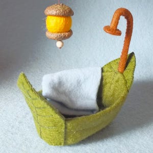 Può includere: Una piccola barca di feltro con una coperta blu chiaro all'interno. La barca ha un manico in feltro marrone e una luce in feltro giallo appesa sopra.