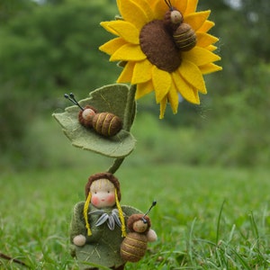 Puede incluir: Una muñeca de fieltro con cabello amarillo y un vestido verde se encuentra frente a un girasol de fieltro con tres abejas de fieltro. La muñeca sostiene una abeja de fieltro en su mano.