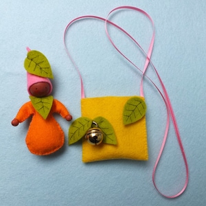 Op de afbeelding: Een kleine vilten pop met oranje en roze kleding en groene bladeren, die een hoed met groene bladeren draagt. De pop houdt een klein geel vilten tasje vast met een groen blad en een bel.