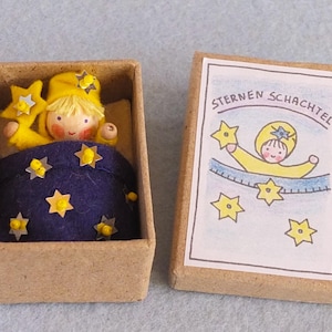 Könnte beinhalten: Eine kleine Holzpuppe in Gelb mit einer blauen Decke befindet sich in einer braunen Schachtel. Der Schachteldeckel hat eine Zeichnung eines Kindes in gelber Kleidung mit Sternen und dem Text "Sternen Schachtel".