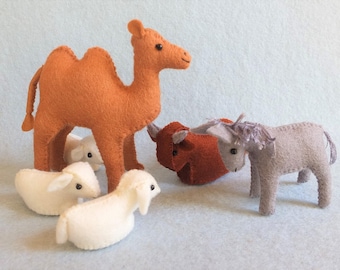 May include: A felt nativity scene featuring a large orange camel, a red ox, a gray donkey, and two white sheep. The handmade figures are arranged on a light blue surface.