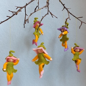 Op de afbeelding: Vijf vilten bloemenfeeën die aan een tak hangen. Elke fee heeft een roze bloemenhoofd, een groen lichaam en gele benen. De feeën hangen met een touwtje aan een tak.