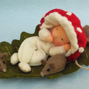 May include: A handmade felt doll, dressed in a white onesie and a red mushroom hat, sleeps on a green felt leaf with three brown felt mice.