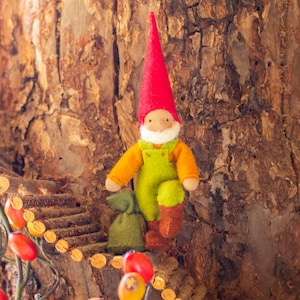 May include: A felt gnome doll with a red hat, green overalls, and brown boots is climbing a wooden ladder made of twigs. The gnome is holding a small green bag. The ladder is attached to a tree trunk with brown bark.