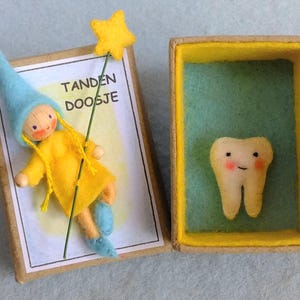 Puede incluir: Una pequeña muñeca con un sombrero azul y ropa amarilla sostiene un diente en una pequeña caja de madera. La caja está etiquetada como "Tanden Doosje".