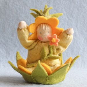 May include: A felt doll with a yellow flower head and green leaves, wearing a yellow sweater and holding a pink flower. The doll is sitting in a green and yellow felt flower.