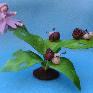 May include: A purple felt fairy doll with wings sits on a green felt leaf with three brown felt snails. The snails have black felt antennae.